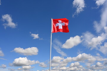 The swiss flag flutters in the wind in a blue sky with white clouds