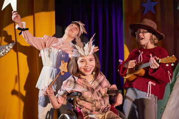 Portrait of happy young girl using wheelchair acting out enchantress on stage holding wooden magic stick in hand, while performing with group of fellow actors at drama school for children