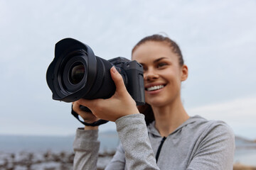 Obraz premium Young woman smiling while holding a professional camera by the beach, showcasing a passion for photography and a joyful connection with nature