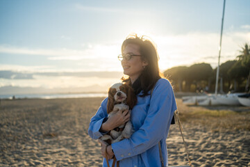 A young woman hugs her Cavalier King Charles Spaniel on the beach at sunset. Warm light, waves, and...