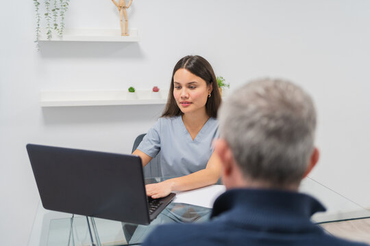 Young physiotherapist using laptop while talking with senior patient in her modern and bright medical office