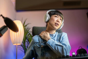 Beautiful Asian female gamer with neck pain during live streaming. Neon lighting, painful facial...