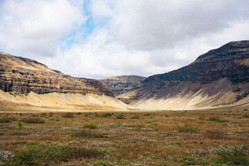 Wide valley between layered volcanic mountains in Iceland, under a cloudy sky with patches of blue,...
