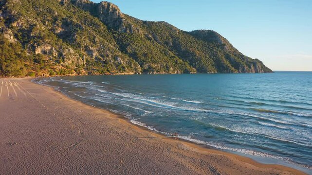 As a stunning sunset unfolds on Iztuzu Beach, the drone slowly pans to follow a woman walking along the shore. Lengthening shadows, golden lights, and gently lapping waves.