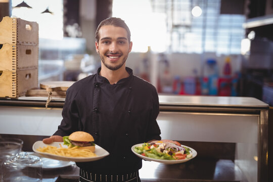 Male chef presenting two plates of food in restaurant kitchen, showcasing burger fries and salad