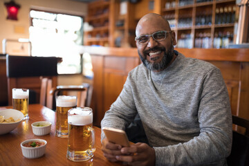 Indian man enjoying beer and chips with salsa at wooden pub table, smiling and using smartphone