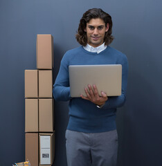 Middle Eastern young adult man holding silver laptop standing in office space with stacked boxes