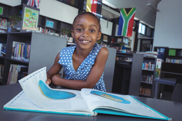 African American girl leaning on study table in public library, smiling at camera with open atlas