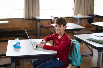 Young boy typing on silver laptop in school classroom, with blue pencil case and teal backpack