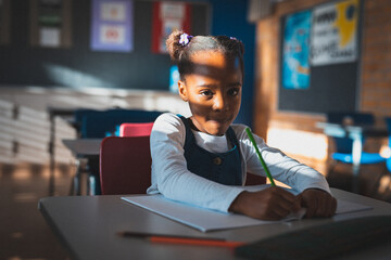 African American girl sitting at desk in school classroom, writing with green pencil and notebook