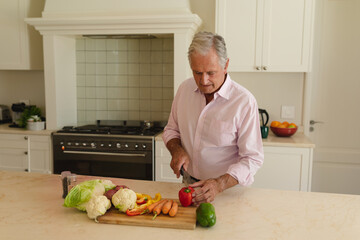 Senior man slicing red bell pepper on wooden cutting board in modern kitchen, with fresh vegetables