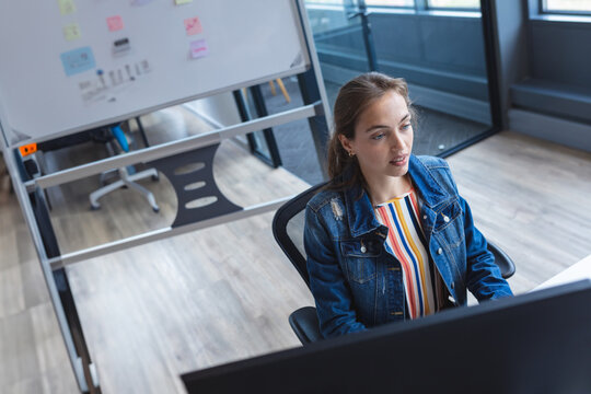 Woman analyzing data on computer monitor while sitting at modern office desk, with whiteboard stand - Powered by Adobe