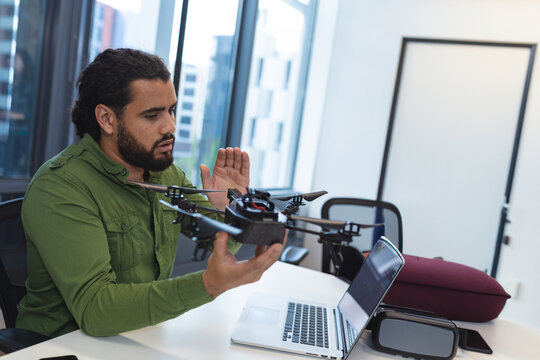 Man inspecting quadcopter drone at modern office desk by window, featuring laptop and VR headset