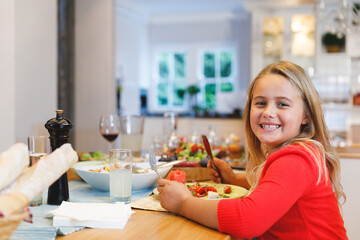 Seven-year-old girl sitting at dining table in kitchen, with salad bowl, bread rolls, copy space