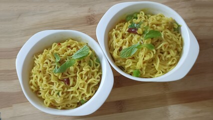Two bowls of yellow instant noodles mixed with green peas and chili, topped with fresh mint leaves. Served in white square bowls on a light wooden surface under natural light.
