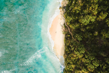 Drone shot of a secluded beach in Bali, with crystal-clear water, crashing waves, and dense tropical vegetation along the rugged coastline.