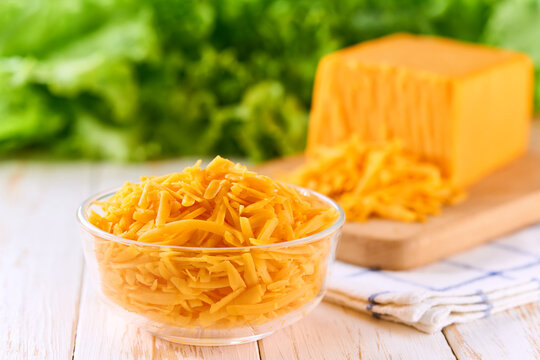 grated cheddar cheese in a clear glass bowl on a white wooden table, selective focus.