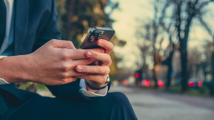 Close up Male hands with smartphone, outdoors. Businessman is typing text messages on mobile phone sitting in the park. Human holds cell phone close up. Man holds mobile phone. Connection concept.