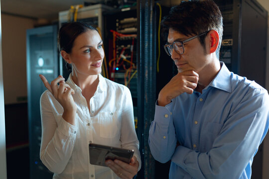 Diverse coworkers examining server racks and cables with blinking LEDs in server room, using tablet