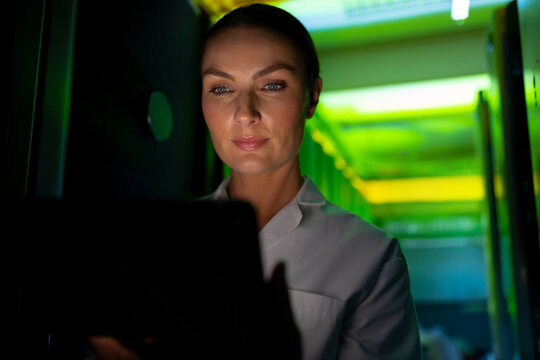Woman checking tablet in data center corridor, surrounded by server racks and LED lights