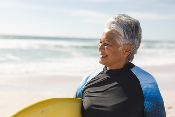 Senior woman standing with yellow surfboard on beach in black or blue wetsuit, enjoying ocean breeze