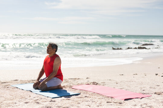 Senior man kneeling on blue yoga mat at sandy beach near shoreline rocks, copy space
