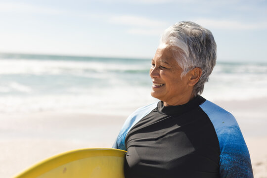 Senior woman standing with yellow surfboard on beach in black or blue wetsuit, enjoying ocean breeze