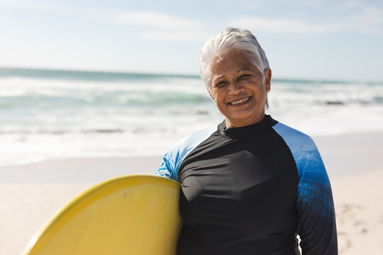 Senior woman standing at sandy beach water's edge holding yellow surfboard, smiling under clear sky