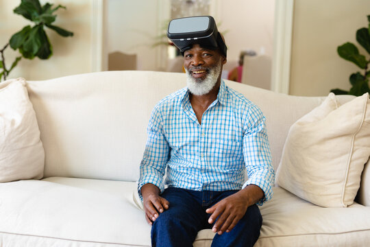 Senior African American man sitting on sofa with pillows in living room, pushing VR headset upward