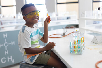 African American boy wearing goggles holding orange test tube in lab with tube rack, copy space
