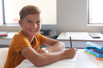 9-year-old boy writing in open notebook at classroom desk with blue pencil case, smiling