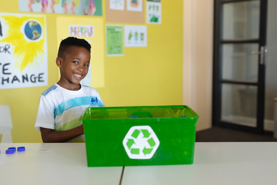 African American boy placing clear plastic bottle into green recycling bin in classroom, copy space