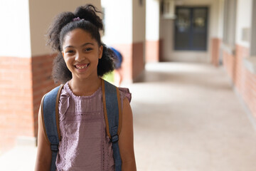 Female child student standing in school walkway with brick columns, windows, wearing blue backpack