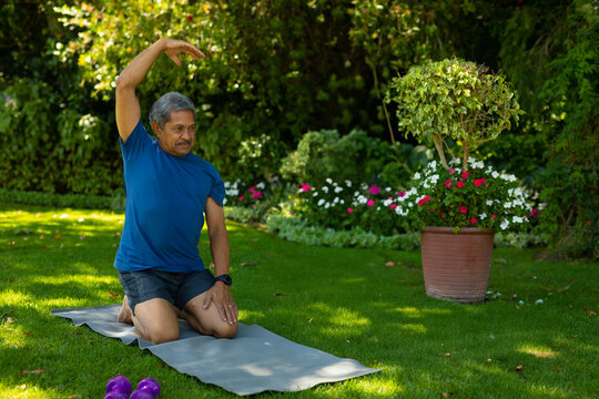 Senior man kneeling, stretching side in backyard, with purple dumbbells terracotta pot, copy space - Powered by Adobe