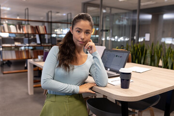 Fototapeta premium Asian woman leaning against standing desk in modern open-plan office, with laptop and coffee cup