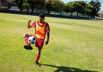 Young man juggling soccer ball on grass field in daylight, wearing red uniform and orange vest
