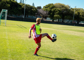 Male soccer player controlling soccer ball on grass soccer field, with goalpost and fluorescent bib