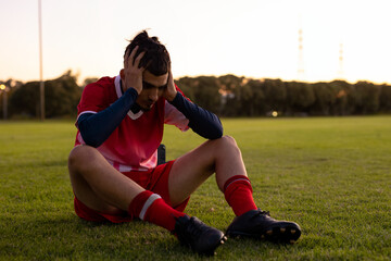 Young adult male soccer player sitting on grass at sunset, holding head in hands, wearing cleats