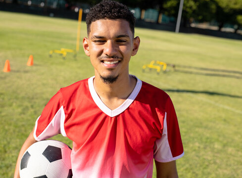 Male soccer player practicing drill on outdoor training field, holding ball with cones and hurdles