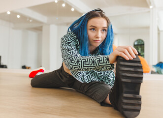 Non-binary person stretching leg wide on dance studio floor, reaching boot sole near studio mirror