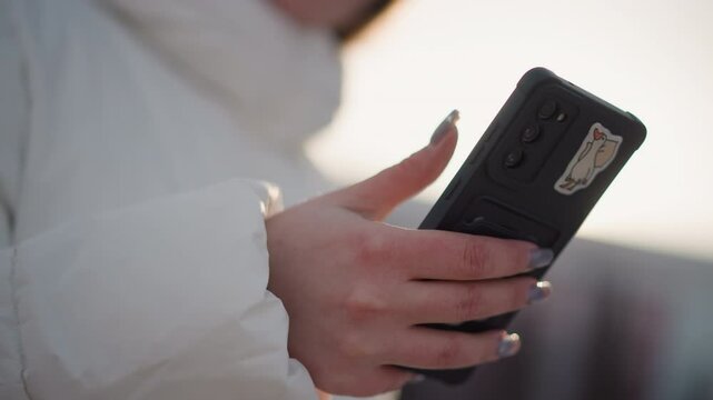 Smart phone with black case held by manicured hand scrolling through apps as young creative strolls through snowy park wearing winter coat headphones and tinted goggles under glowing sunlight