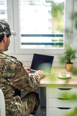 Fototapeta premium vertical Soldier working from home using laptop and wearing camouflage uniform