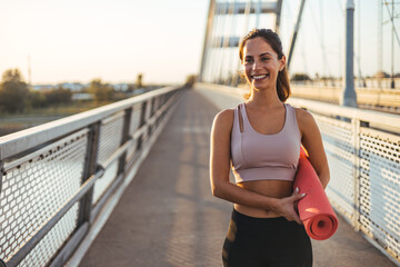 Smiling Woman Holding Yoga Mat on Bridge During Sunrise