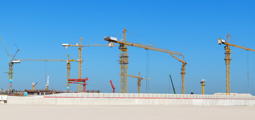 Multiple tower cranes at a large construction site under a clear blue sky. The image highlights the scale and intensity of modern construction projects with heavy machinery and structural growth