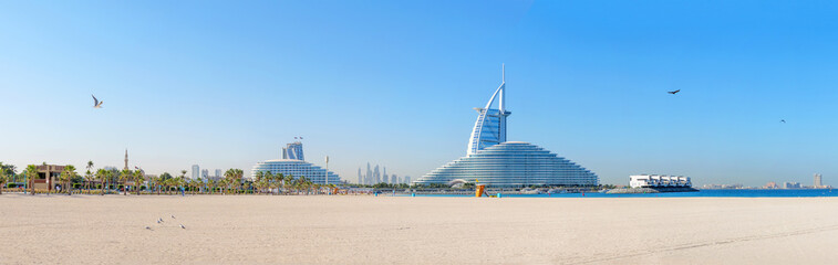 Fototapeta premium Panoramic view of Dubai city with sandy beach, clear blue sky, and palm trees. Dubai skyline in the background. A perfect sunny day in Dubai, ideal for tourism and travel scenes
