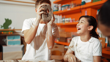 Smart boy pick clay while looking though hole at pottery workshop.Skilled diverse highschool student working and modeling dough while laugh at happy child at art lesson. Creative art. Edification.