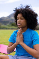 African American boy meditating on pink yoga mat in grassy field with rolling hills