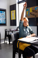 Girl child raising hand in modern classroom, holding pen over open notebook on desk