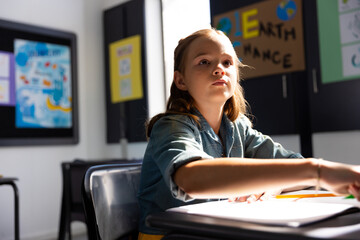 Eight year old girl writing at desk in classroom with screen and Earth poster, copy space