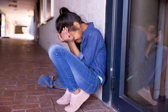 Asian child girl sitting in school corridor hugging knees and crying with blue backpack, copy space